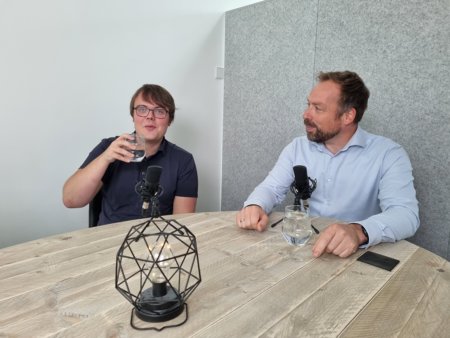 Foto van opname podcast datagedreven sociaal domein, links Robin Hoogervorst van Datogether, rechts Aad Francissen van 8ting, beide zittend aan een ovale houten tafel met een glas water en een microfoon.