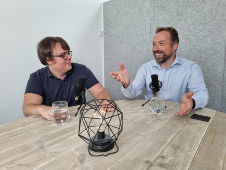 Foto van opname podcast datagedreven sociaal domein in de studio aan de Moutlaan 20, Enschede. Links Robin Hoogervorst van Datogether kijkt enigzins verbaast naar rechts. Daar zit Aad Francissen van 8ting, die enthousiast en lachend met handgebaren iets vertelt. Beide zitten aan een ovale houten tafel met een glas water en een microfoon.